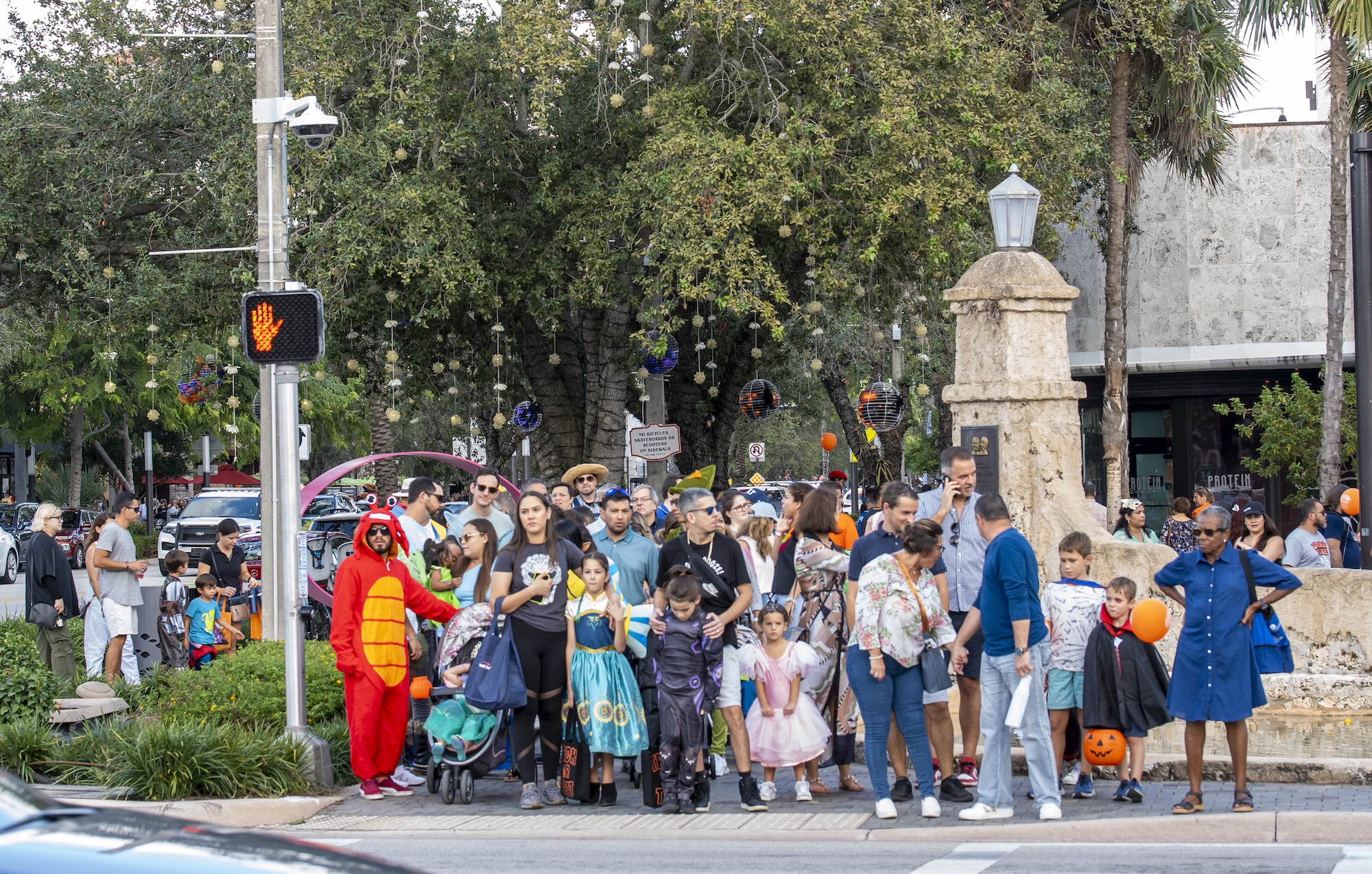 a big group of trick-or-treaters in costume and their parents wait to cross the street at a crosswalk