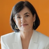 headshot of a woman with shoulder-length dark hair wearing a white blazer against an orange background