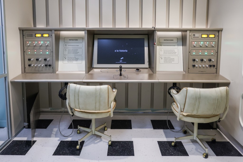 Two vintage plush chairs in front of a board showing a TV and knobs. Two signs, one in English and one in Spanish, ask people to tell their stories to contribute to the Freedom Tower Archive