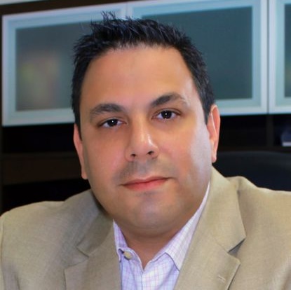 headshot of a short-haired man wearing a beige blazer over a light-colored, open-collared dress shirt