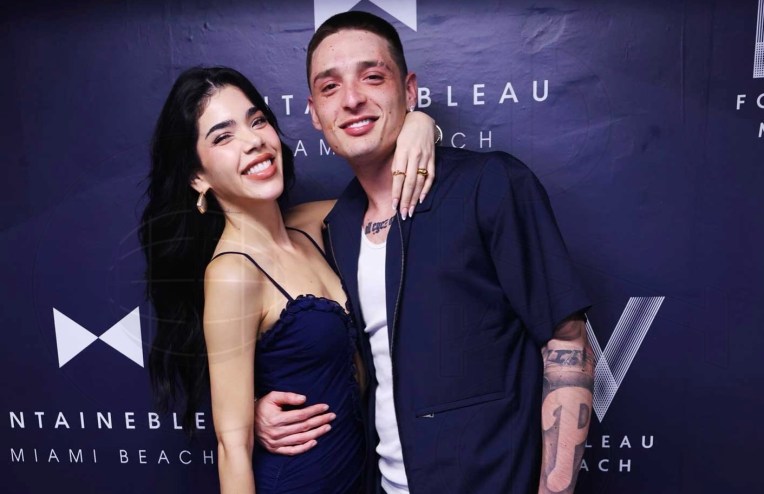 Kenia Os and Peso Pluma hug and pose in front of a step and repeat backdrop reading "Fontainebleau Miami Beach