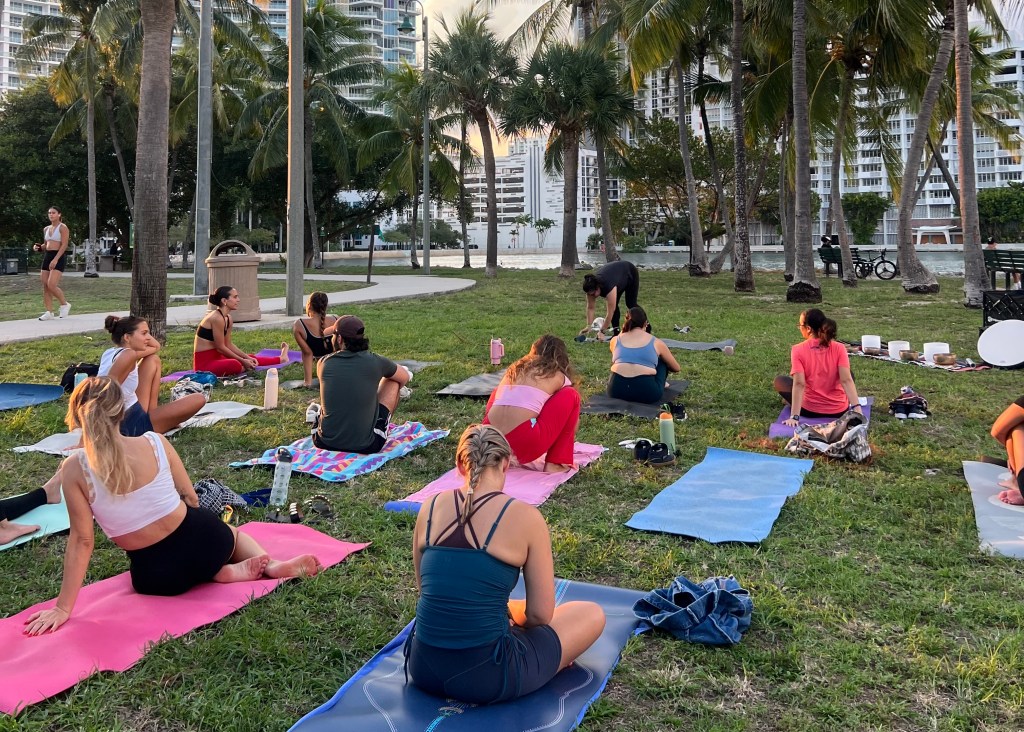a group of mostly women do yoga on the grass in a park in front of the water