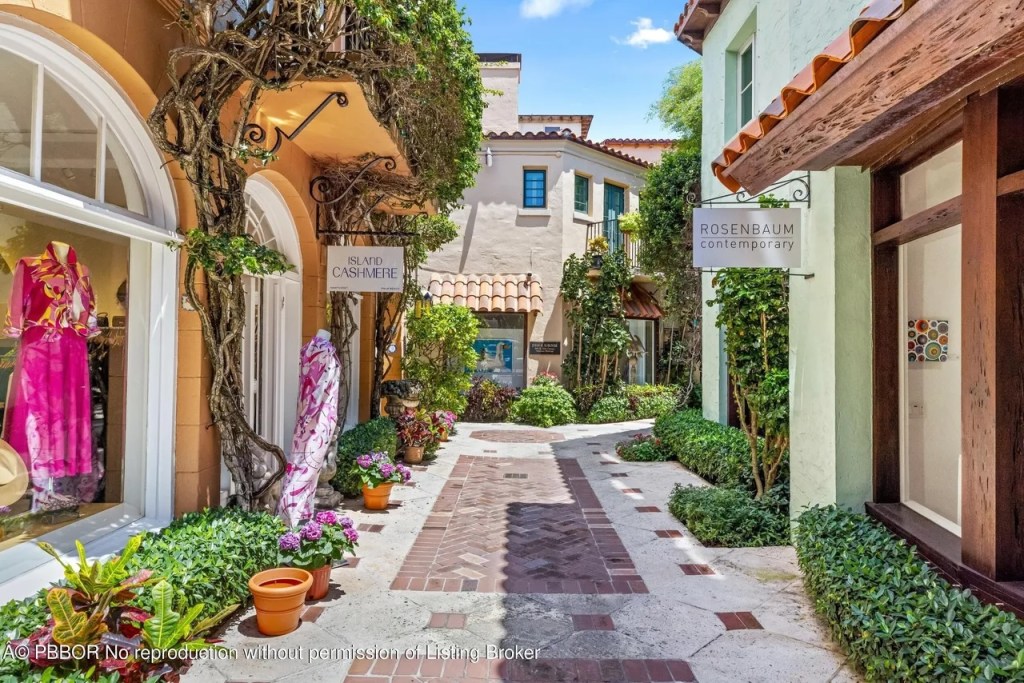 Depicted is a brick path running between retail shops lined with lush South Florida foliage.
