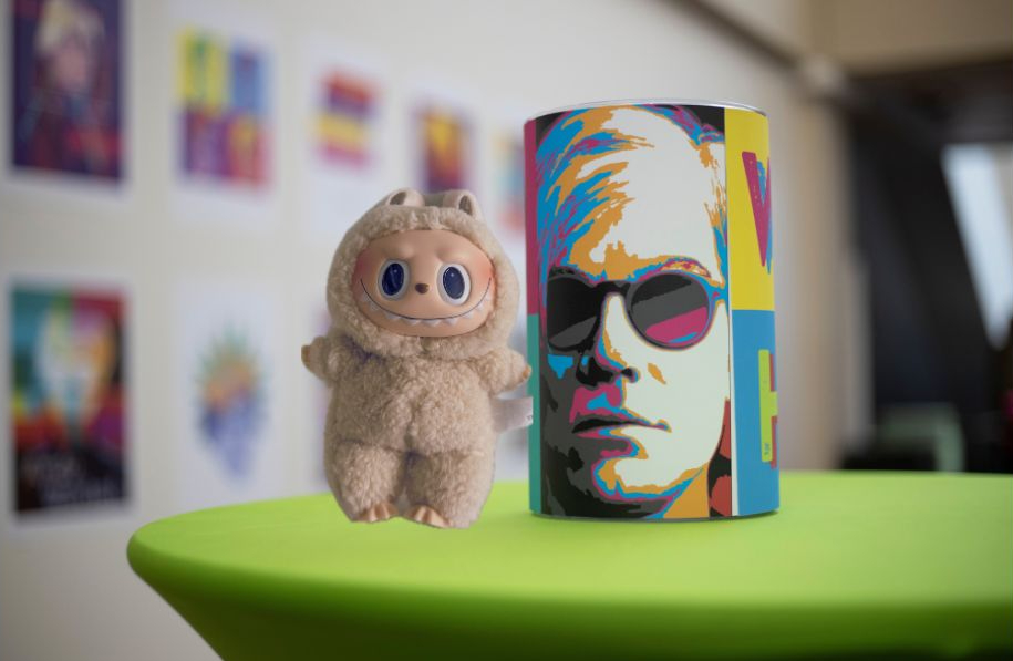 Soymilk Labubu doll standing next to a cup with an image of Andy Warhol. They both are on a green table.