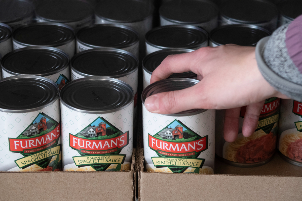 A person grabs a can of spaghetti sauce from a food pantry shelf.