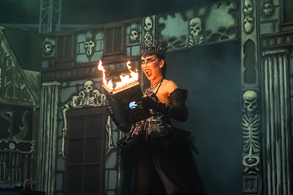 a woman in a black dress and crown and scary makeup holds a book that's on fire