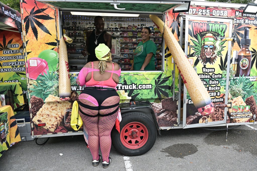 a woman in fishnet clothing stands at the window of a merch truck decorated with inflatable marijuana joints