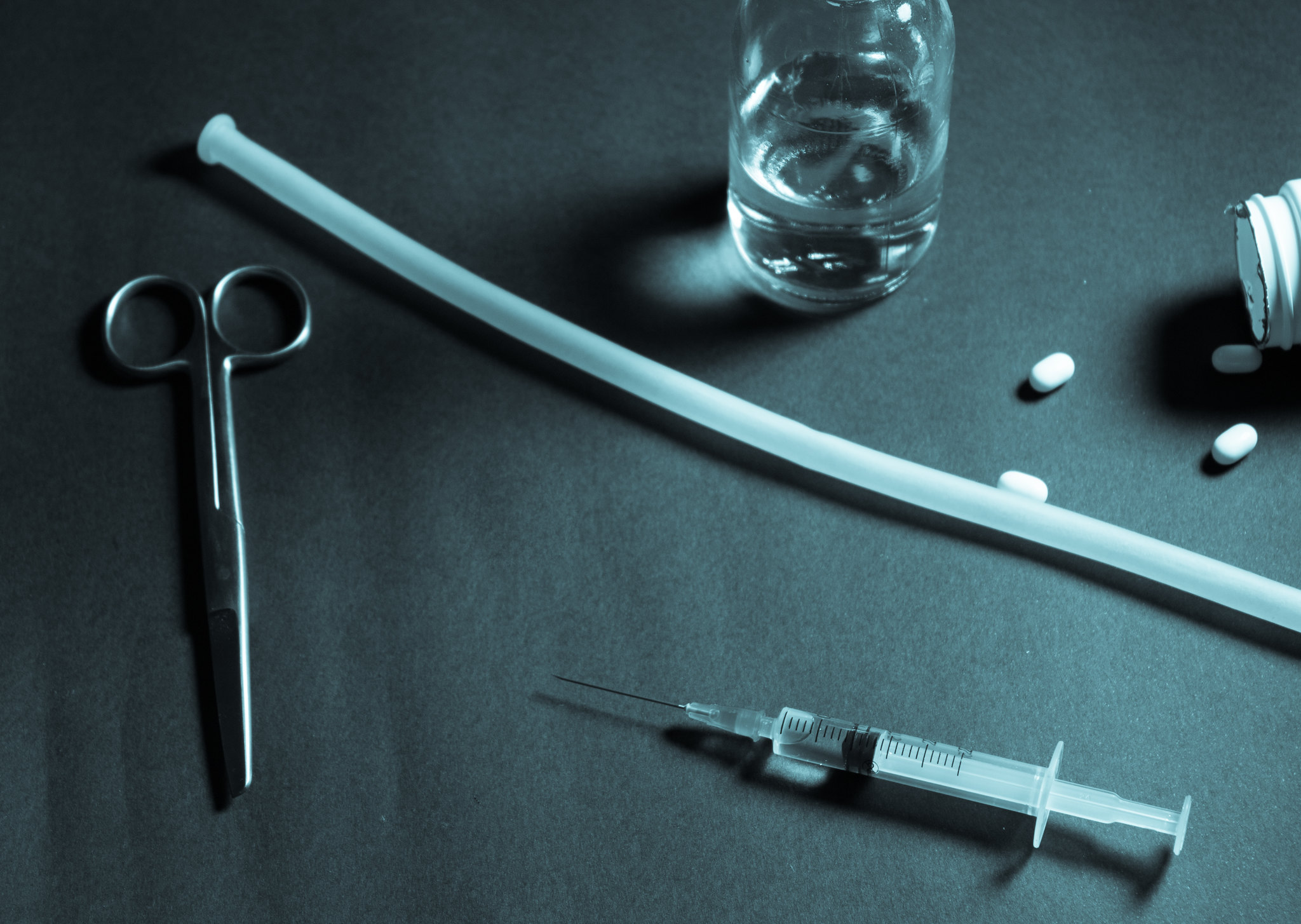 dim photo of a medical tray with instruments including scissors, a plastic tube, a syringe, a glass with clear liquid, and a bottle on its side with three pills spilled out