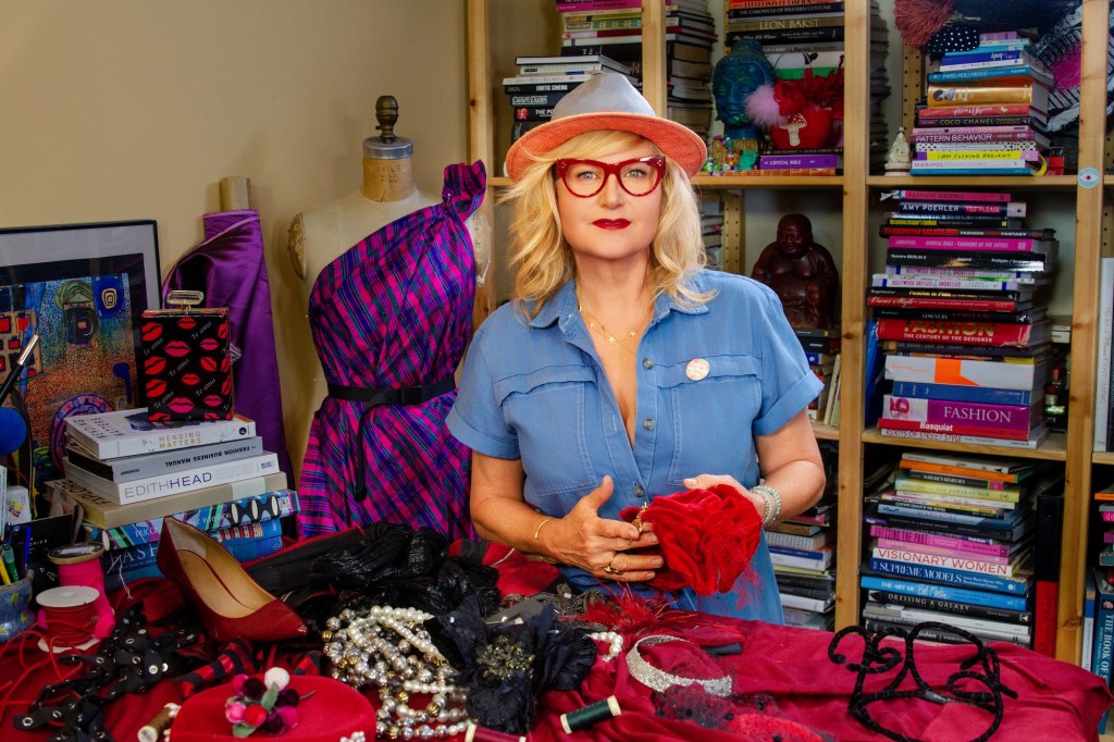 costume designer Mona May in red glasses and a fedora in her studio, surrounded by books, mannequins, and accessories