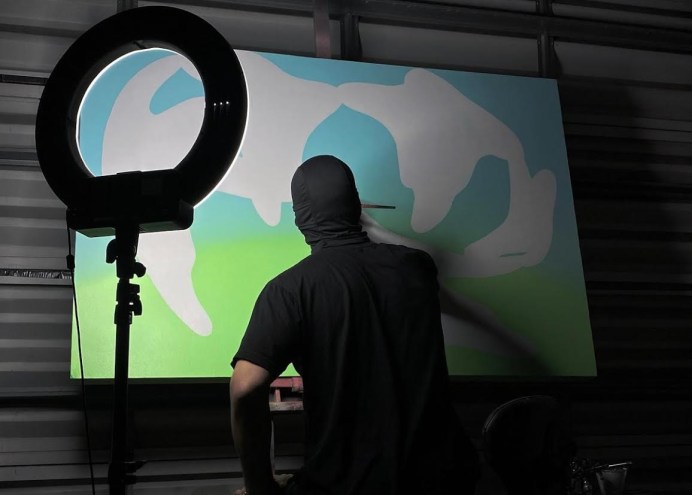 An artist in a black mask obscuring his face and head paints a canvas. A ring light illuminates the canvas.