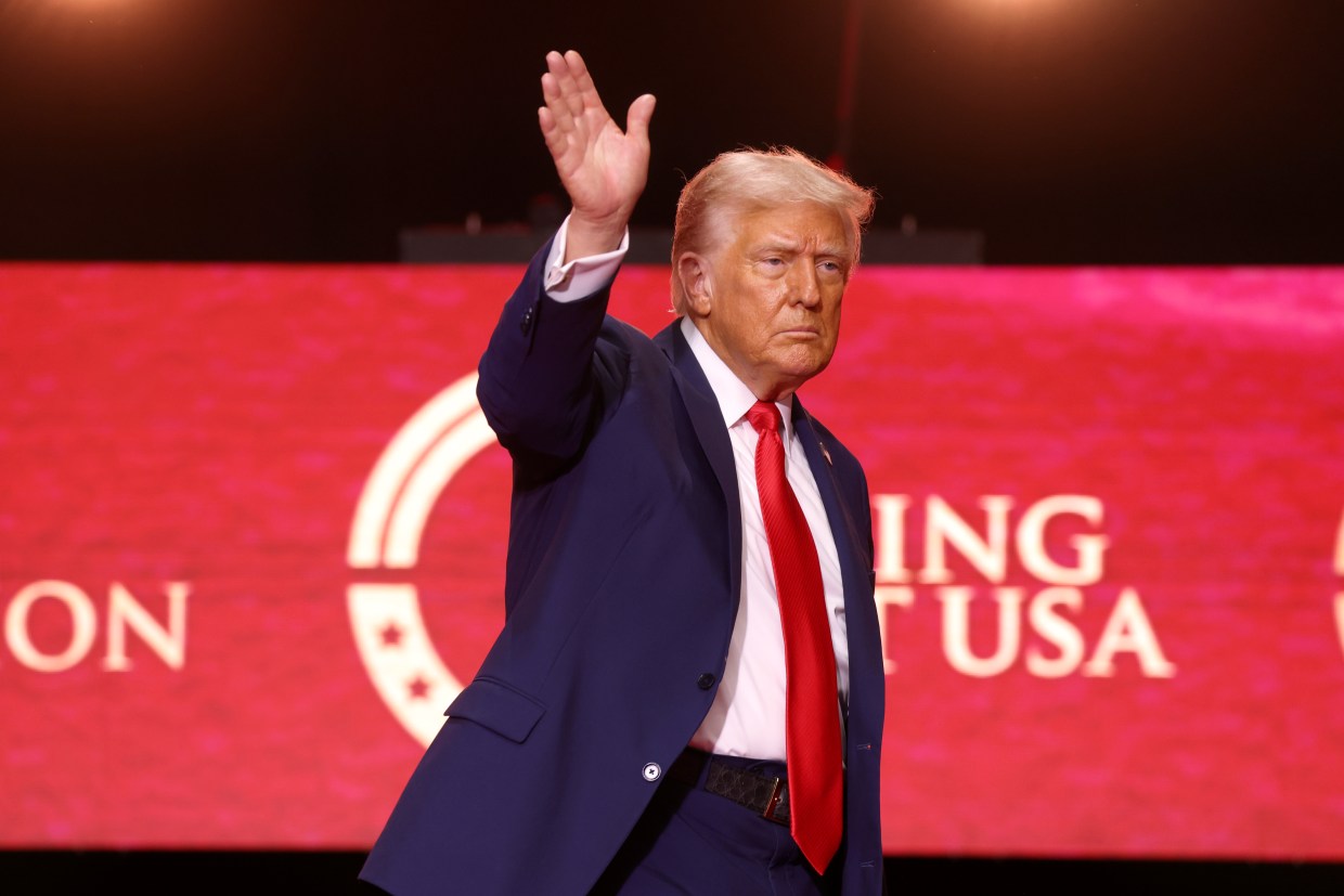 Donald Trump waving his right hand. He is wearing a red tie and standing in front of Turning Point USA red banner.