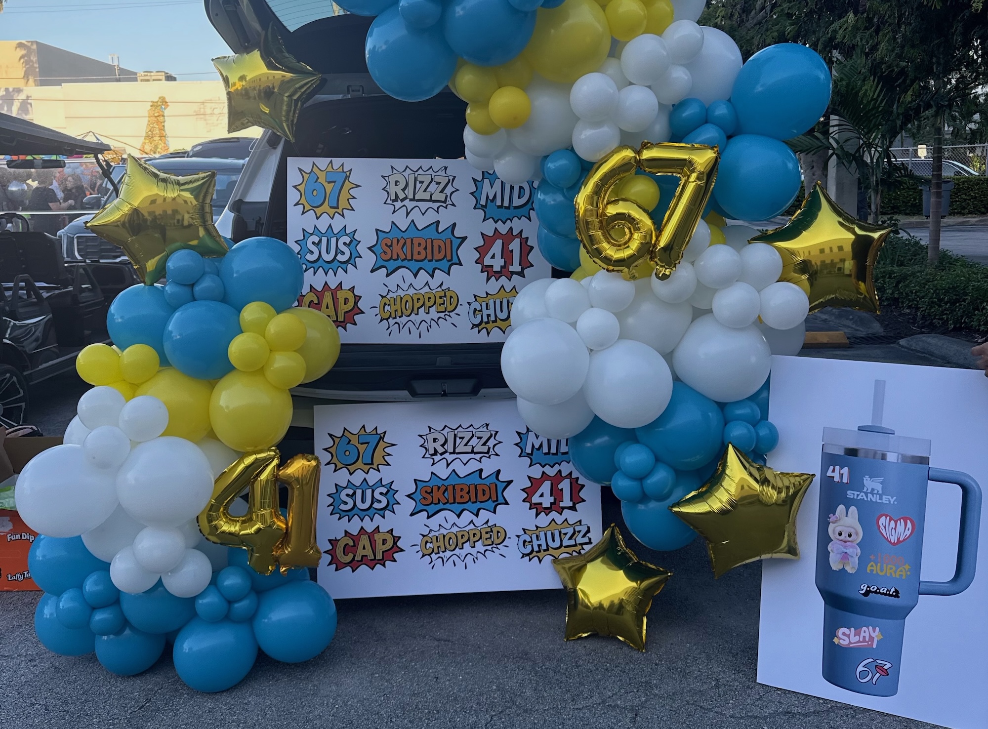 a photo of an open SUV trunk with white, blue, and gold balloons and posters with nonsense phrases like "67," "skibidi," and "rizz"
