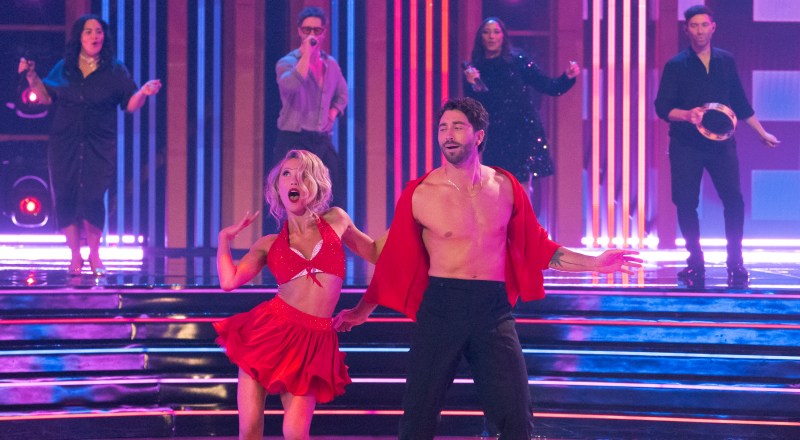Photo of a woman and man dancing on a stage. The woman is on the left wearing a bedazzled red bralette and skirt with a bedazzled white bra underneath. The man, on the right, wears black pants and an open red, silk shirt. Behind them are two female singers, one male singer, and a man playing tambourine