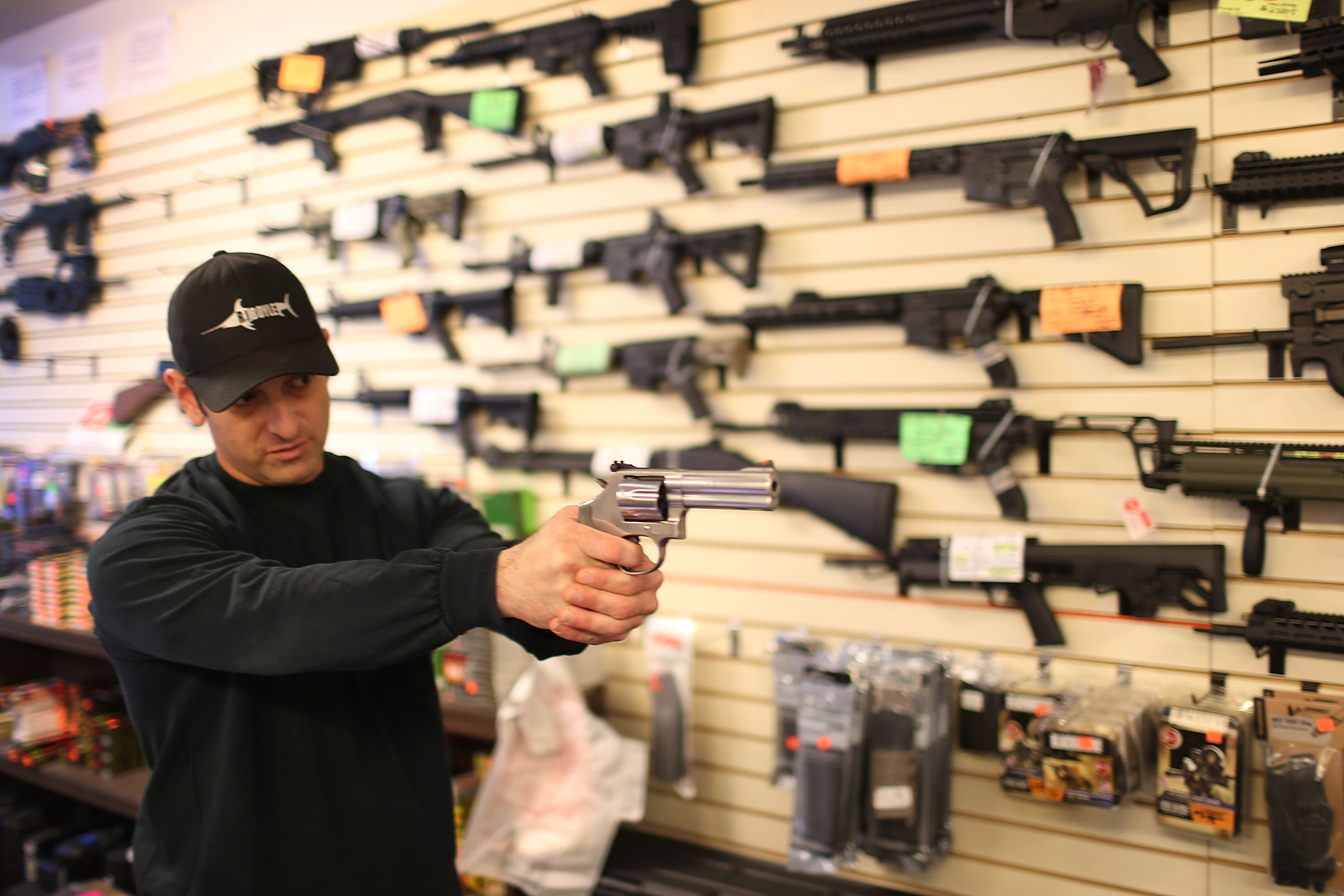 a gun shop employee sights down a handgun against a backdrop of a wall filled with semiautomatic rifles