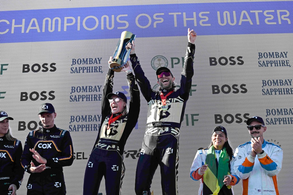 Photo of three two-person racing teams in racing suits. The duo in the middle holds up a trophy underneath a sign reading, "Champions of the Water"