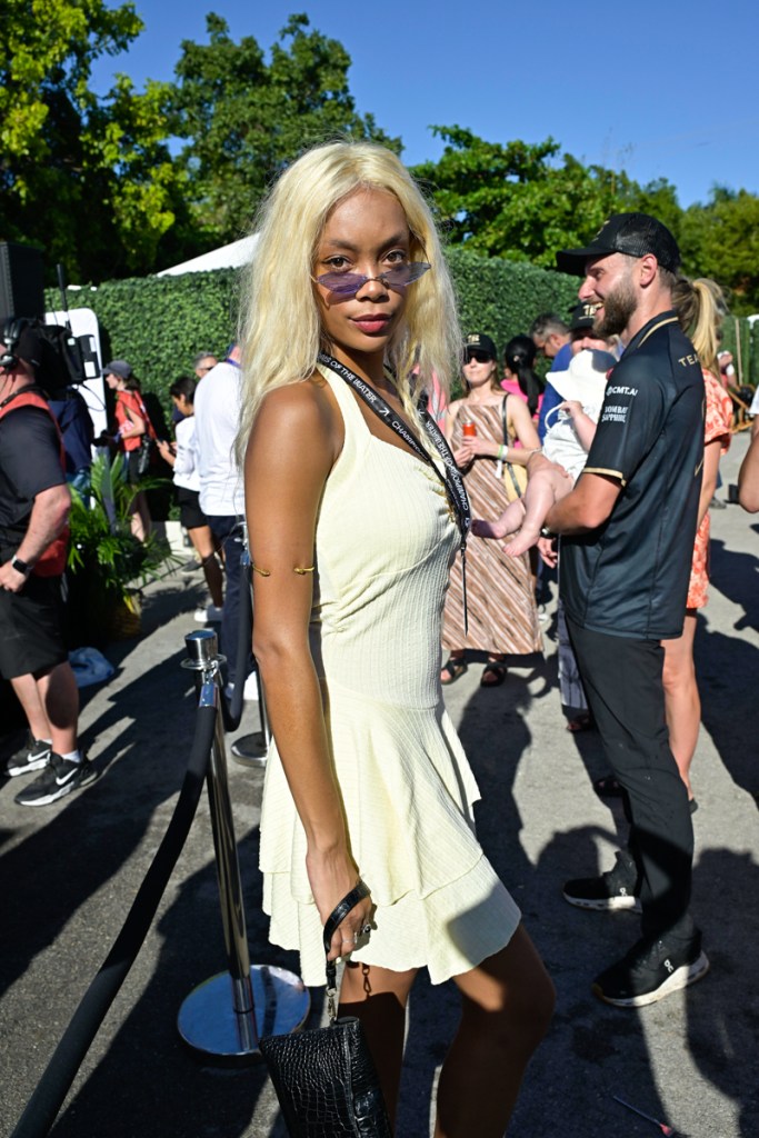 photo of a woman in geometric shades and a pastel dress wearing a lanyard and holding a black purse as she poses in front of a crowd at an outdoor sporting event