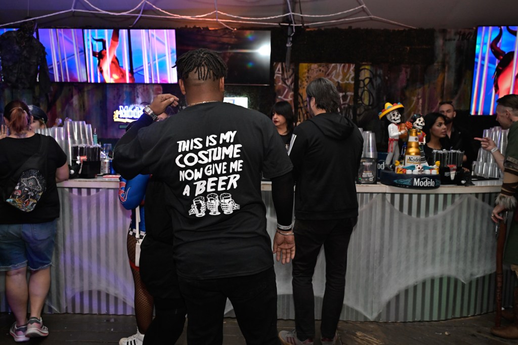 Photo of people waiting at in line at a bar at a Halloween party. The back of the t-shirt of the main person in the photo reads, "This is my costume now give me beer"
