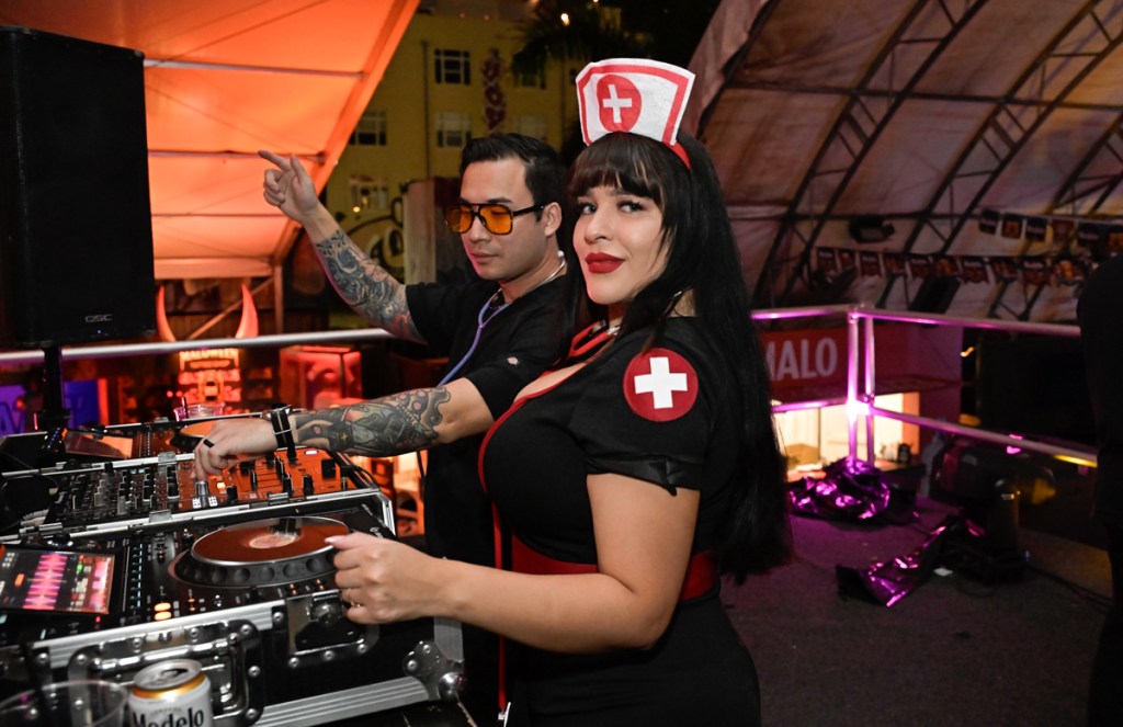Photo of two people behind a DJ booth on Halloween. The woman is dressed as a nurse