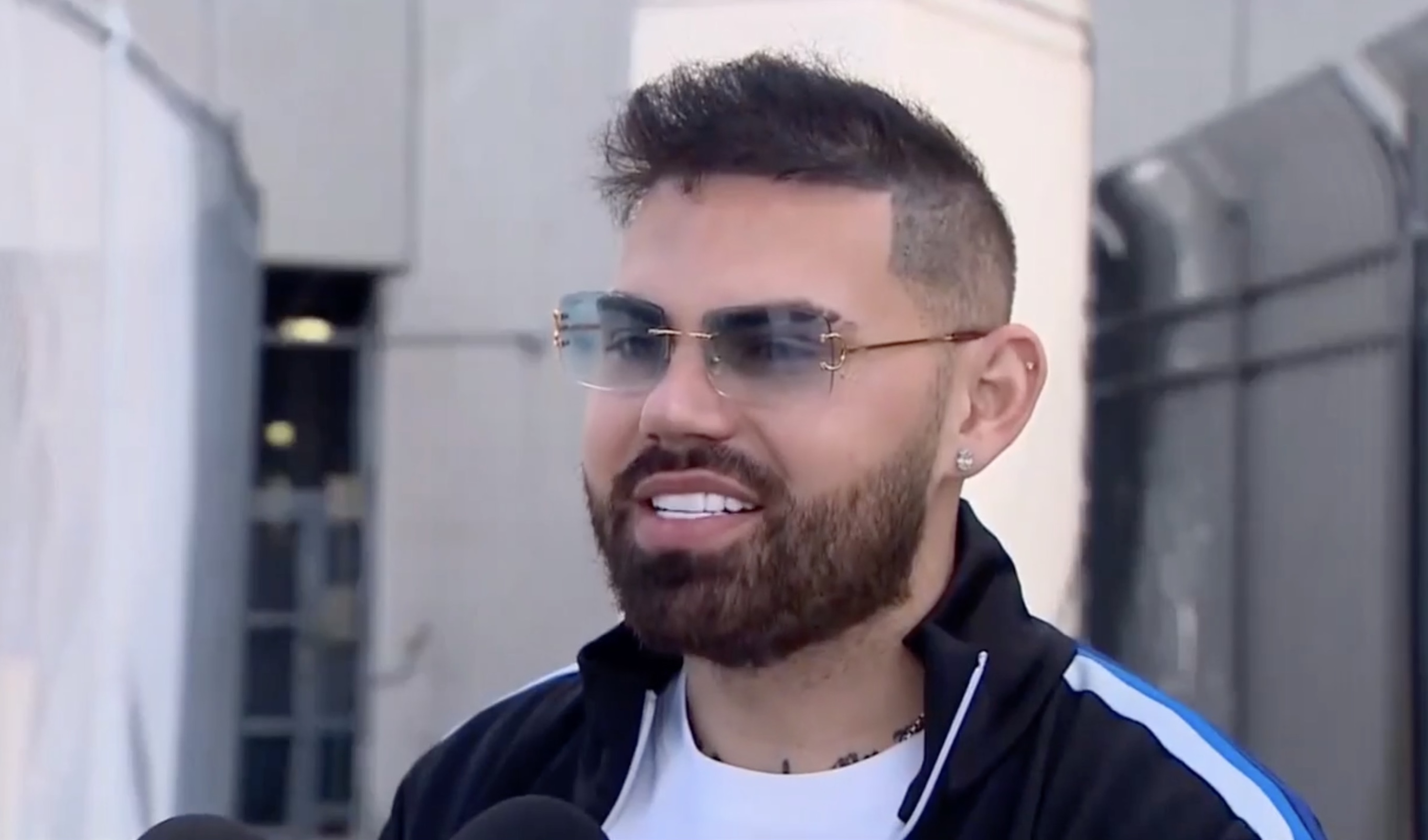 TV news screenshot of a casually dressed bearded man with a fade haircut, shown head to shoulders wearing tinted frameless glasses and appearing to speak to reporters