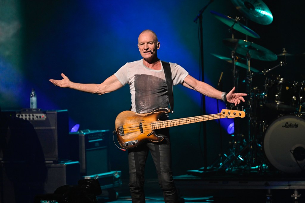 Picture of Sting holing a bass on stage with his arms open. 