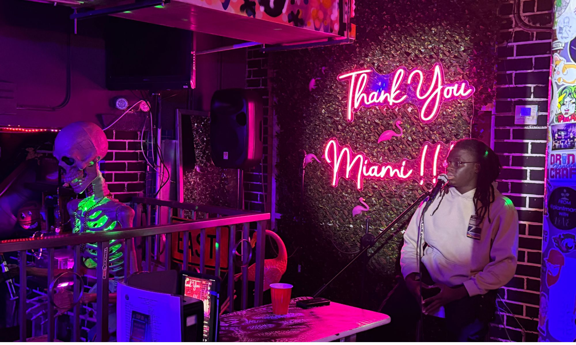 Photo of a neon sign that reads Thank you Miami and a person talking on a microphone.