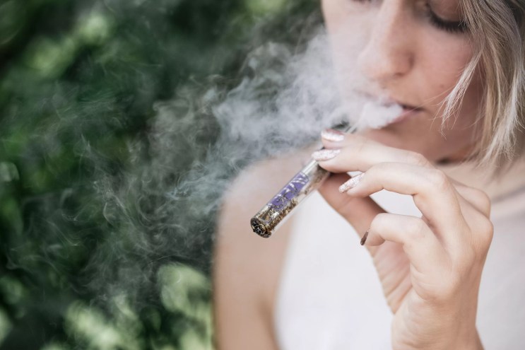 A woman with short hair who is wearing a white tank top puffs on a one hitter pipe packed with what appears to be marijuana while sitting outdoors.