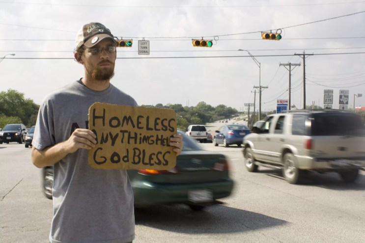 A man who appears to be homeless stands on the side of a busy road holding a cardboard sign enscribed with the words: "HOMELESS, ANYTHING HELPS, GOD BLESS."