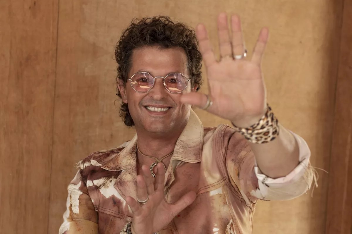 Photo of Carlos Vives wearing glasses and looking at the camera with his left hand up.