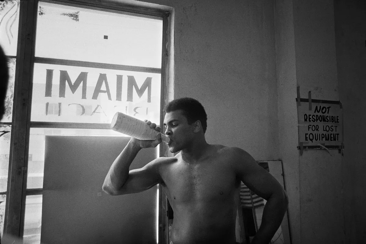 Black and white image of Muhammad Ali in the gym drinking water from a sports bottle