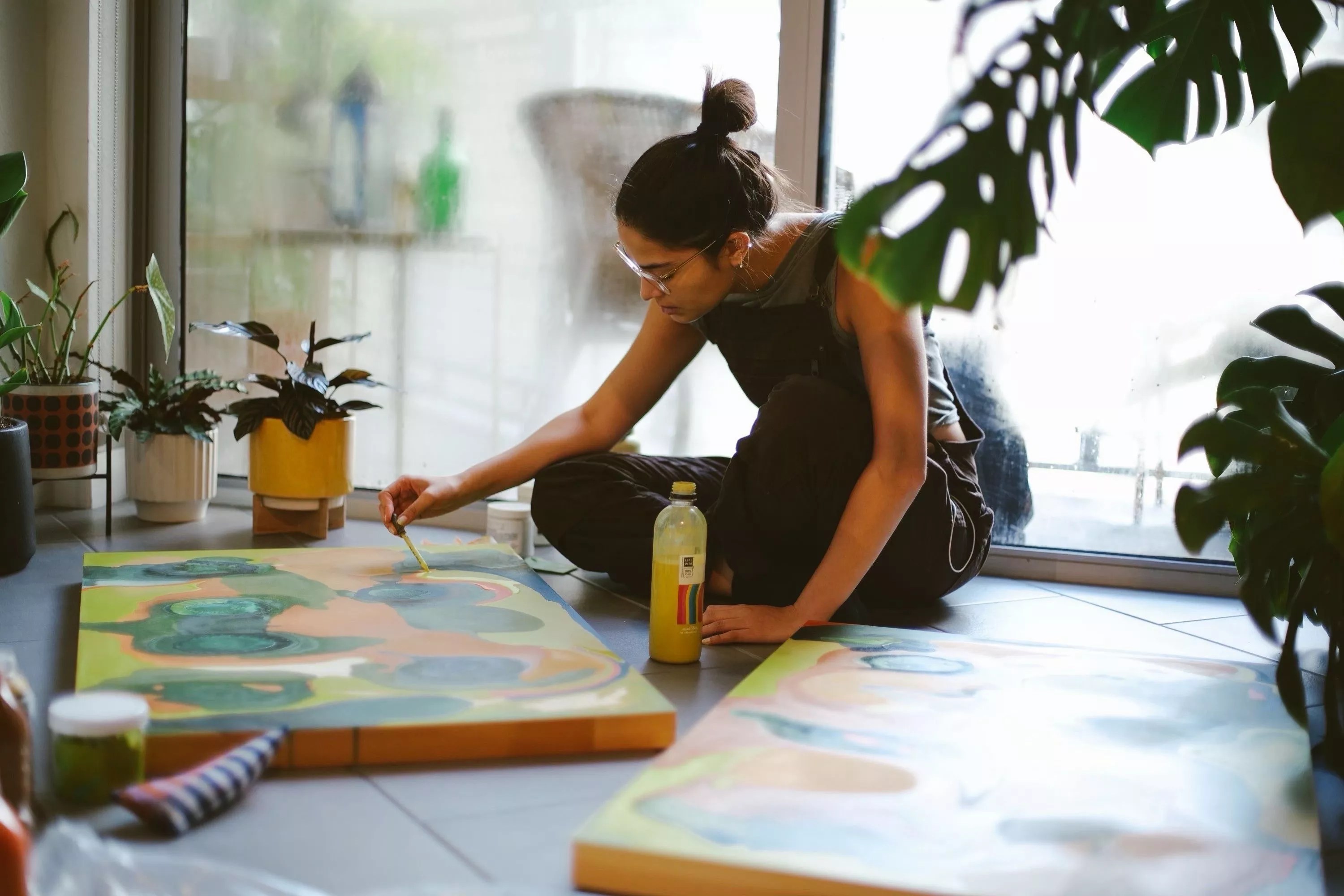 color photo of Miami-based painter Orianna Montenegro at work, seated on the floor of a sunlit room with plants and a large window in the background