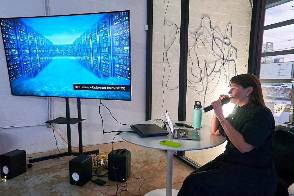 photo of a woman in an art studio speaking into a microphone next to a TV screen showing a video installation