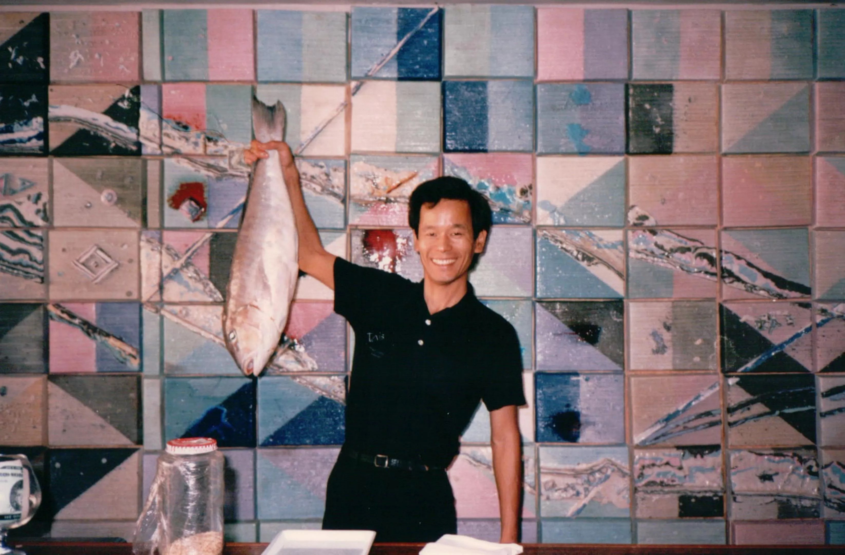 vintage photo of a smiling man dressed in black, standing at a counter against a tiled background and holding up a whole fish in his right hand