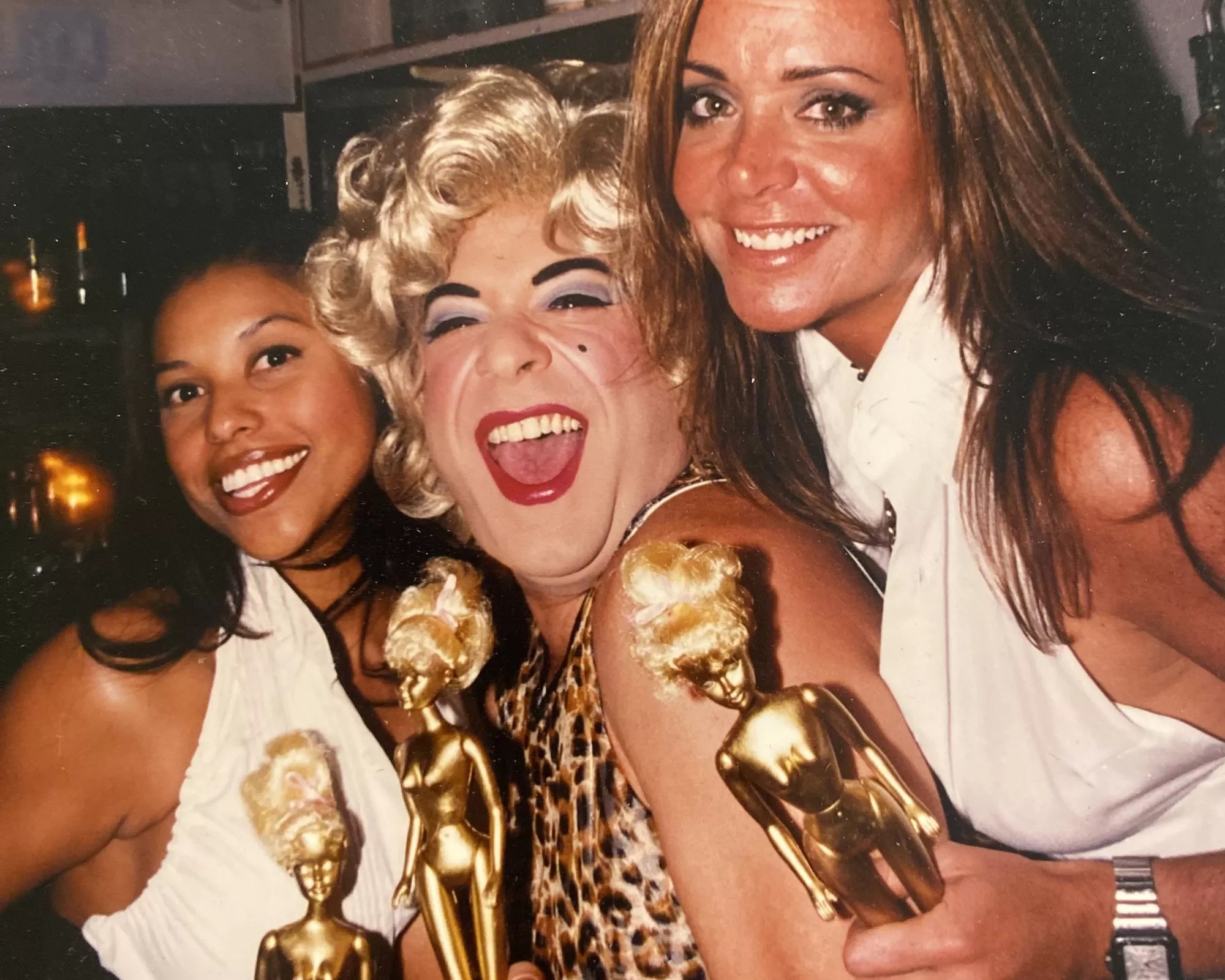 Drag queen Shelley Novak posing alongside two women while holding gold spray-painted Barbies