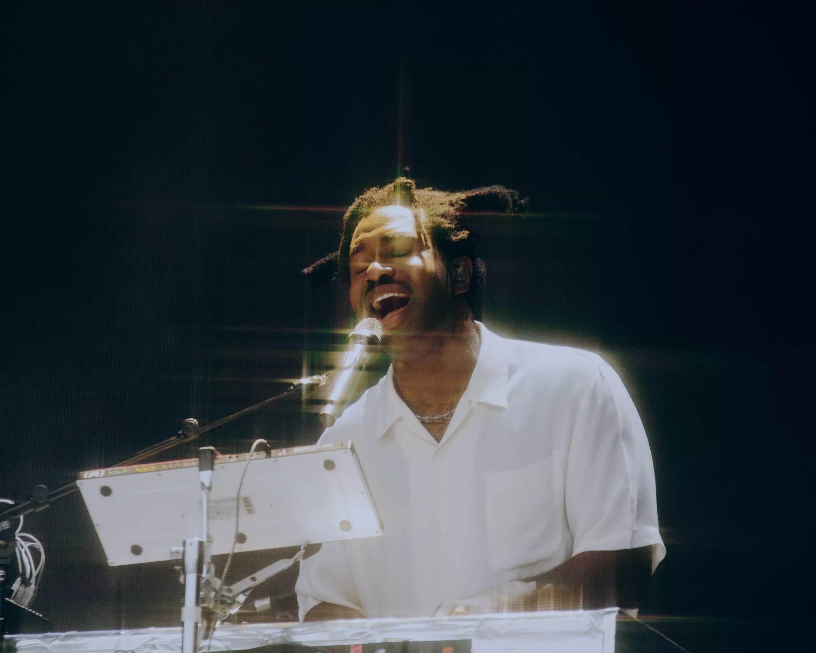 color photo of a white-shirted, dreadlocked Sampha singing into a microphone against a black background