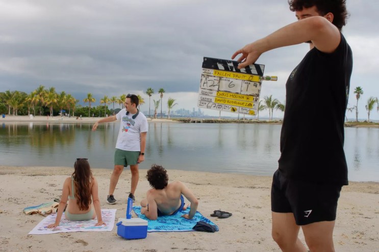 Chris Molina is speaking with actors lying on the beach while a man holds a film slate in the foreground