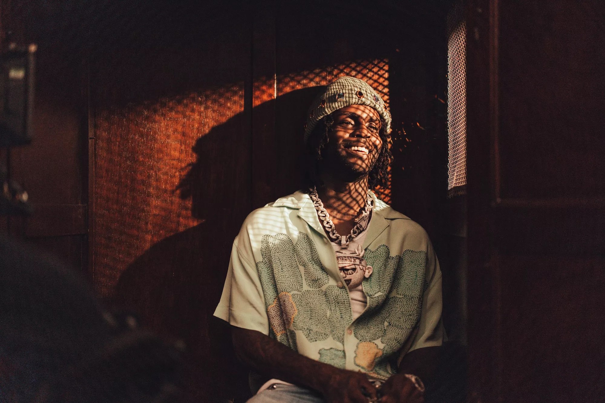 color photo of rapper Chief Keef, smiling, wearing a knit hat and colorful short-sleeved print shirt, and illuminated by sunlight shining through a diamond-patterened mesh screen.