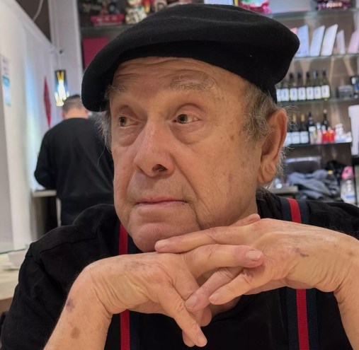 Photo of a 79-year-old man wearing a black beret and a black short-sleeve shirt with red-and-black suspenders, with his hands clasped under his chin.