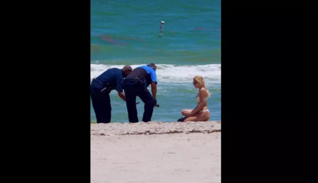 A woman in a bikini and two police officers crouch over a brick of white powder on the beach