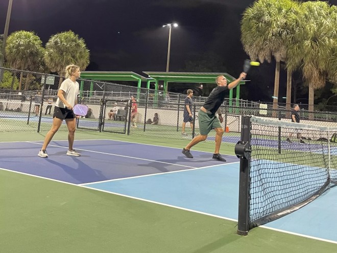 photo of two people playing pickleball on a light blue, navy blue, and green court at night with people sitting and standing in the background behind a fence