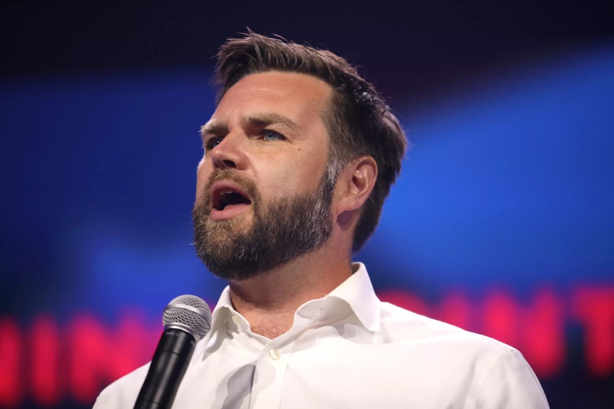 J.D. Vance wearing white open-collared shirt, speaking into a microphone, looking outspoken