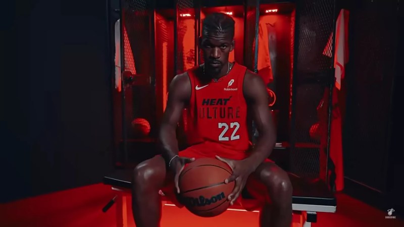 Jimmy Butler wearing the Miami Heat's new blood-red "Heat Culture" jersey, sitting in a dimly lit locker room holding a basketball.