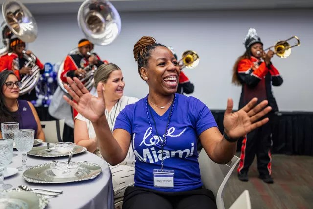 photo of a woman sitting at a banquet table cheering in a shirt that reads, "I love Miami." Two other women sit at the table with her. Two tuba and two trombone players play behind them