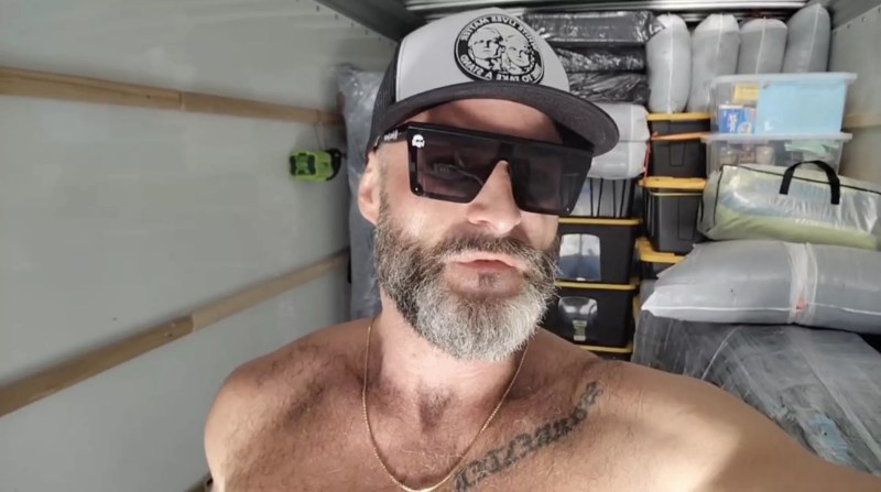 a shirtless Jon Minadeo speaks into the camera inside a loaded U-Haul.