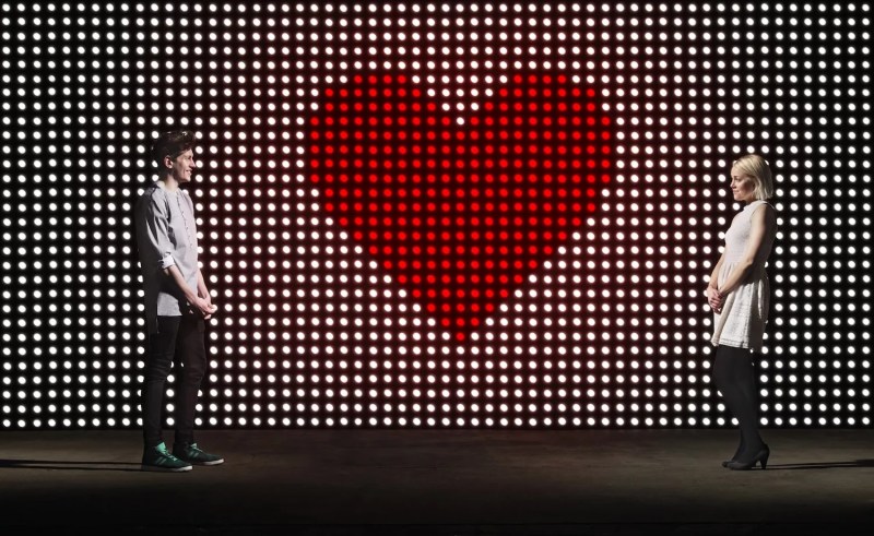 Young couple in front of light wall, with huge heart made of red dots, dating