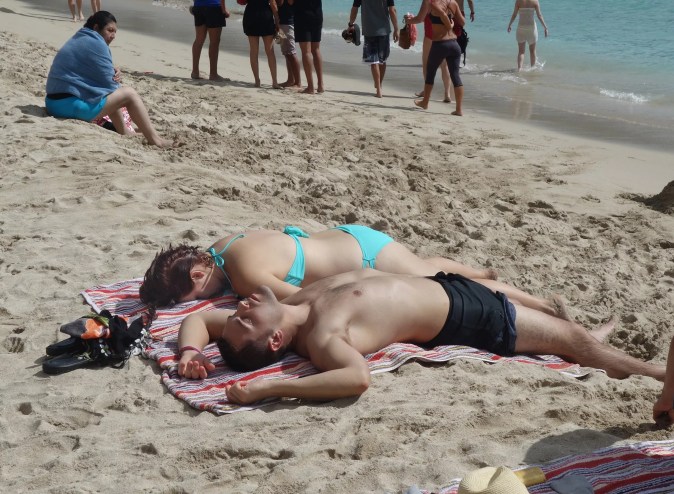 A couple relaxing on sandy shores, lying side by side on a colorful striped towel, soaking up the sun as beachgoers stroll nearby