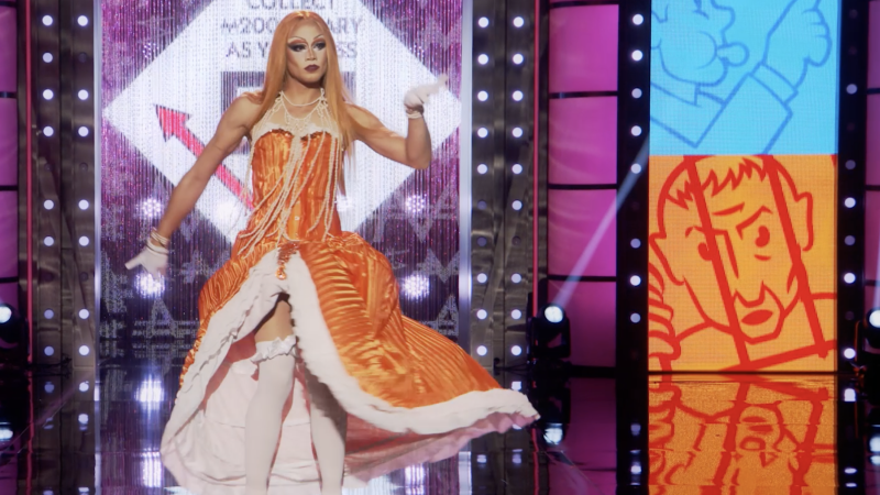 a drag queen walks the runway in an orange wig, orange dress, and flowing pearl necklaces