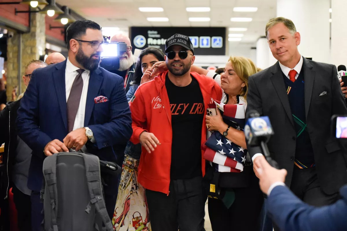 Photo of Enrique Tarrio, in sunglasses, MAGA hat, and red jacket, exiting a Miami International Airport concourse
