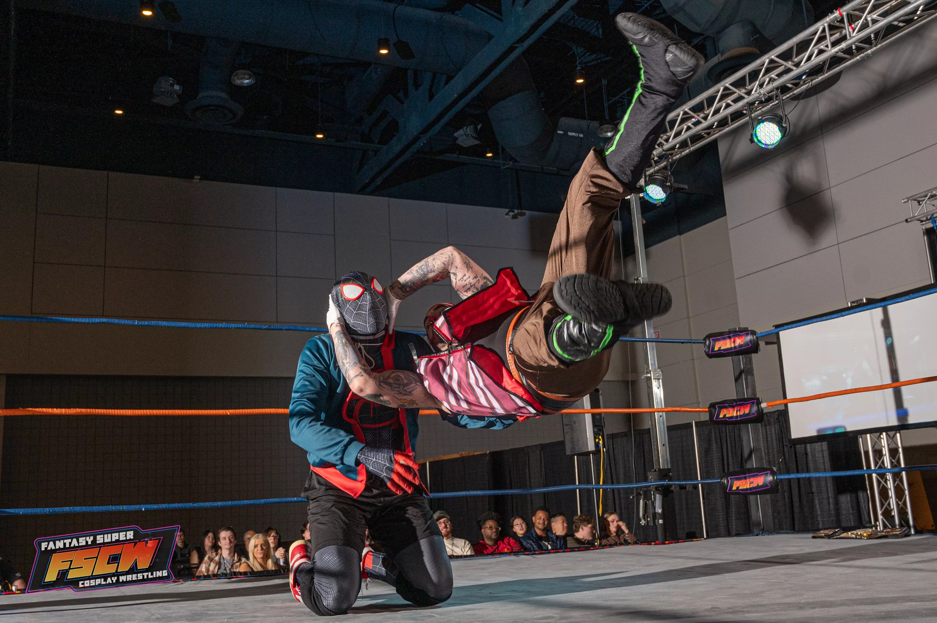 two people dressed in costumes wrestle in a ring. one is suspended in the air parallel to the ground