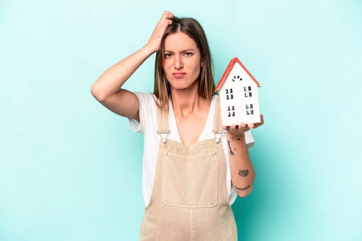 Young white woman holding a toy house and looking peeved, isolated against a blue background