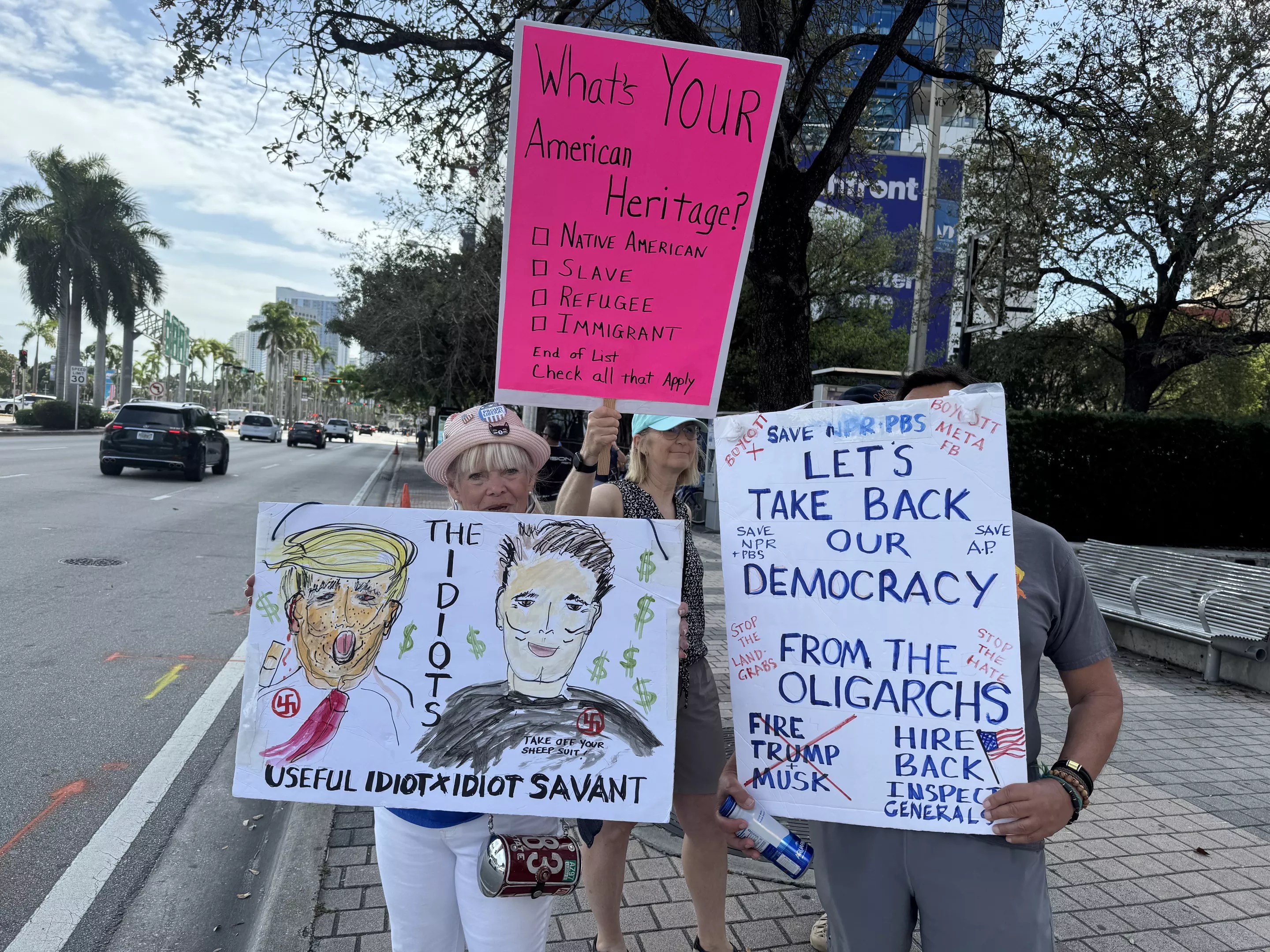 Three Protesters hold their signs that read, "The idiots" with images of Trump and Musk, "Let's take back our democracy," and "What's your American Heritage?"